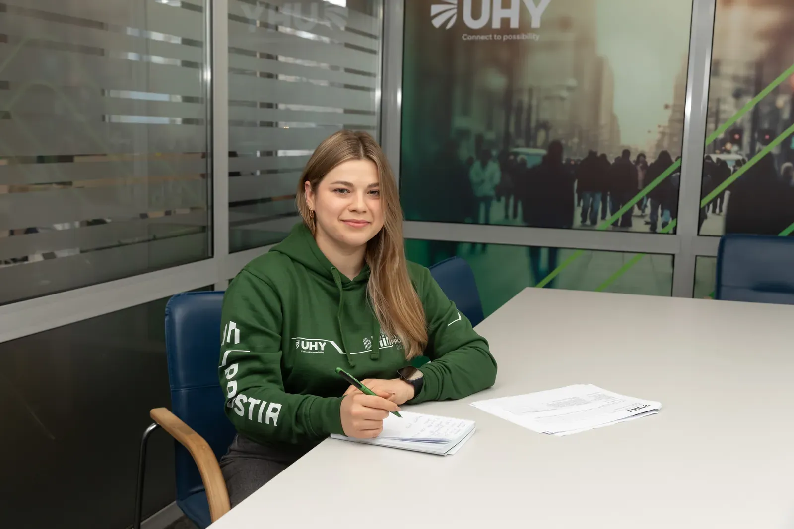 Фахівчиня UHY Prostir у фірмовому худі з документами за столом на тлі банера UHY