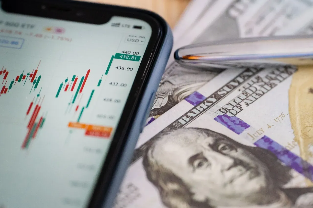 Смартфон с биржевым графиком рядом с долларовыми банкнотами и ручкой